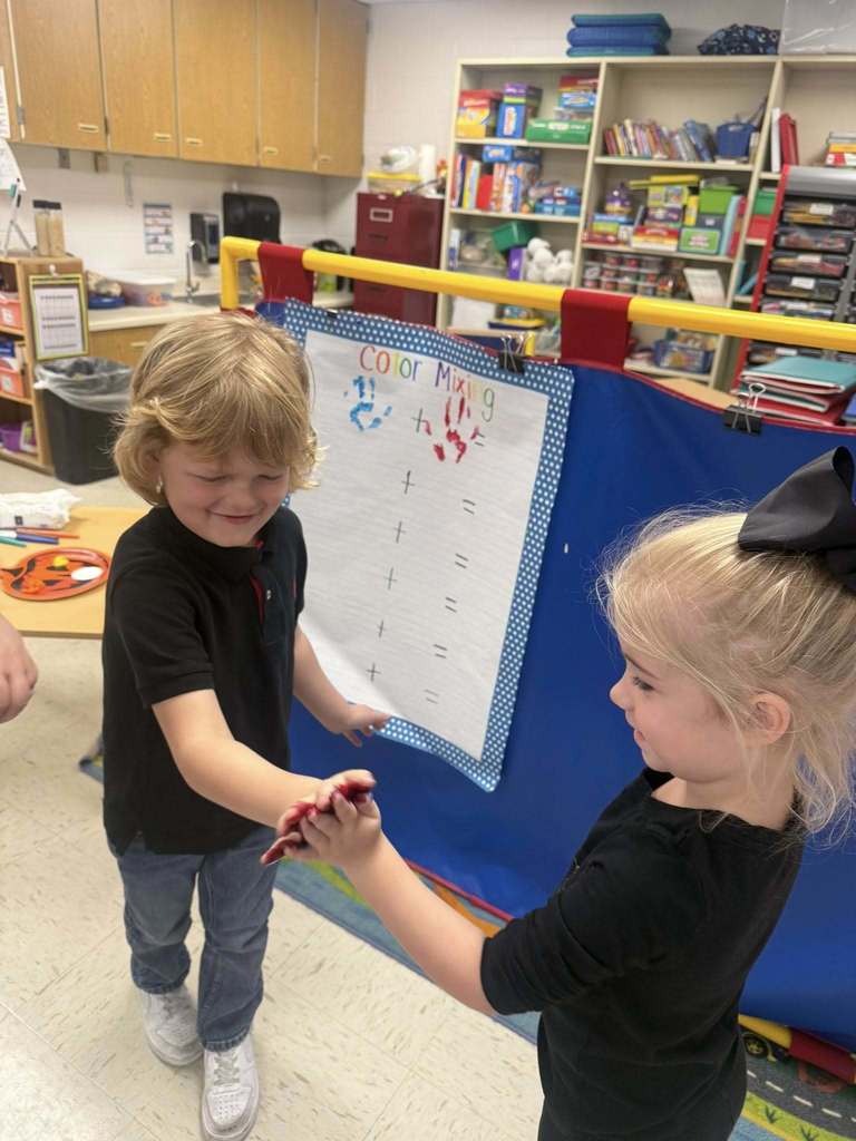 Two Pre-K students press their painted hands together while smiling, with a classroom chart labeled “Color Mixing” displayed behind them.