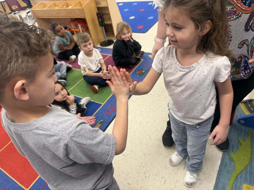 Two Pre-K students press their painted hands together while smiling, with a classroom chart labeled “Color Mixing” displayed behind them.