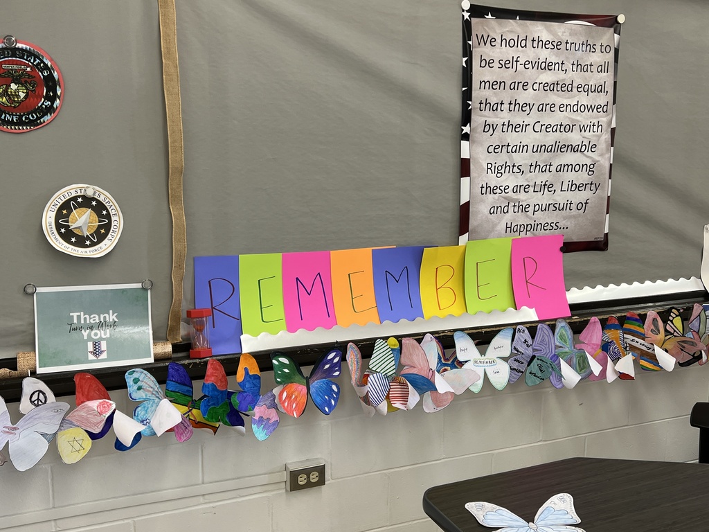  Classroom display with “REMEMBER” letters, student‑decorated butterflies, military emblems, and a Declaration of Independence quote.