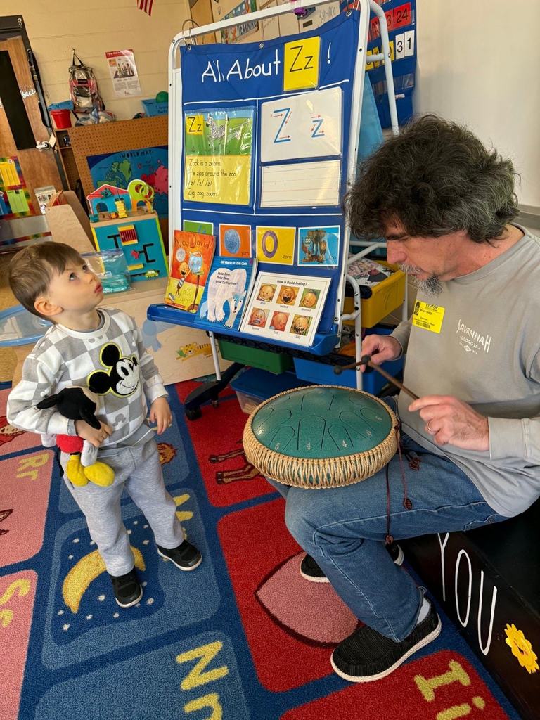 A young student watches closely as a guest musician demonstrates a steel tongue drum, striking the instrument with mallets during a small-group music activity.