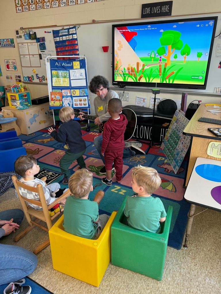 Several Pre-K students gather around a guest musician playing a guitar at the front of the classroom. Some students clap or move while others sit on small chairs and watch.