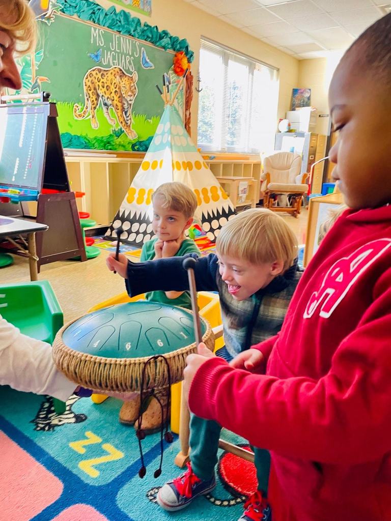 Pre-K students smile and laugh while taking turns striking a steel tongue drum with mallets during a guided music exploration activity.