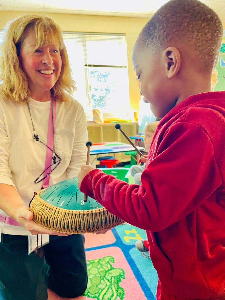 A teacher holds a steel tongue drum steady while a student taps it with mallets, practicing rhythm and coordination during a hands-on music experience.