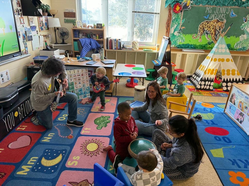 Pre-K students sit and kneel in a classroom circle while a guest musician plays a guitar. Several students hold or tap percussion instruments as a teacher supports the activity nearby.
