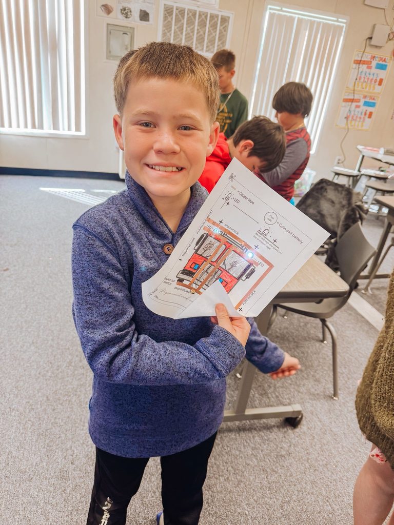 A student proudly holds up a completed paper circuit that lights up, showing copper tape and illuminated components.