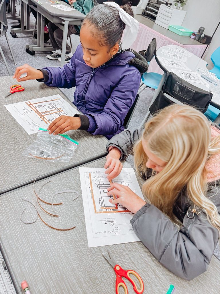 Two students sit side by side assembling paper circuits, aligning copper tape and components on printed templates.