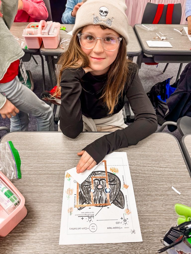 A student smiles while standing next to a completed paper circuit on a desk with visible copper tape pathways.