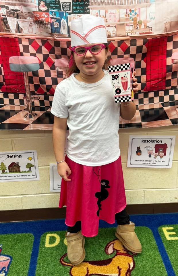 A student smiles while dressed in a 1950s-style outfit and holding a themed cup in front of a classroom display.