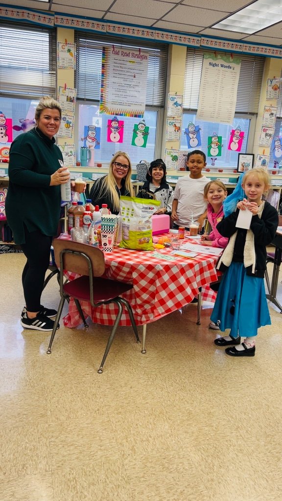 Students and a teacher gather around a table with menus, cups, and play money during a hands-on math and history lesson.