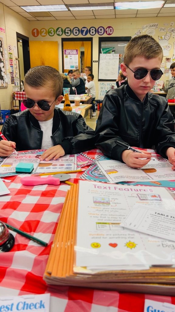 Two students wearing sunglasses complete worksheets at a table set with pencils and guest checks for the themed activity.