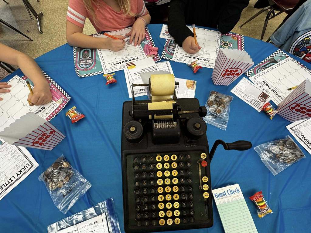 An overhead view shows a vintage-style cash register, guest checks, coins, and worksheets arranged on a classroom table.
