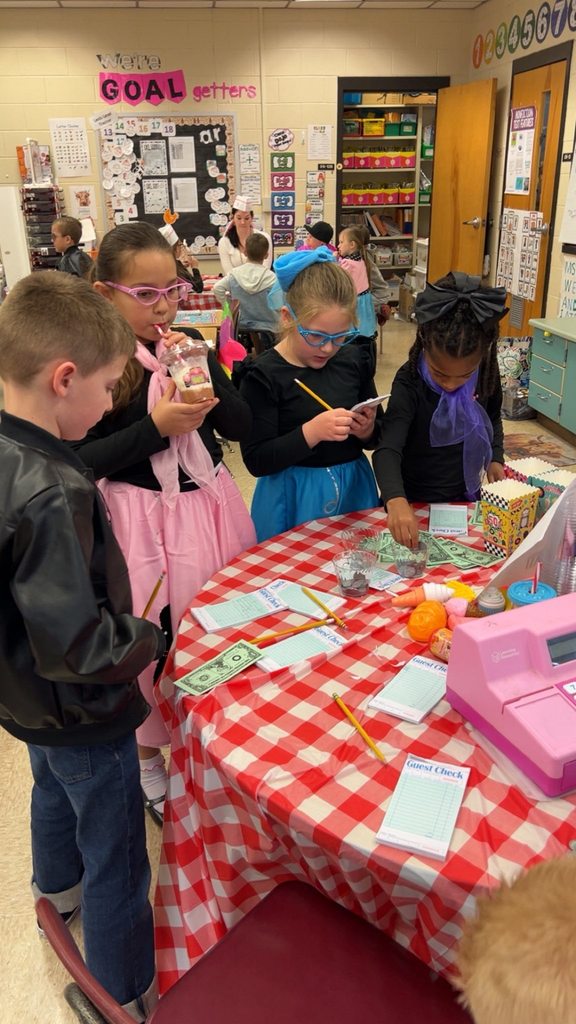 Second grade students stand around a classroom table using menus, guest checks, and play money during a 1950s-themed learning activity.
