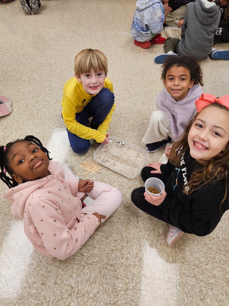Four second grade students sit on the floor around a shallow container of sand and wooden sticks, working together to build a small dam.