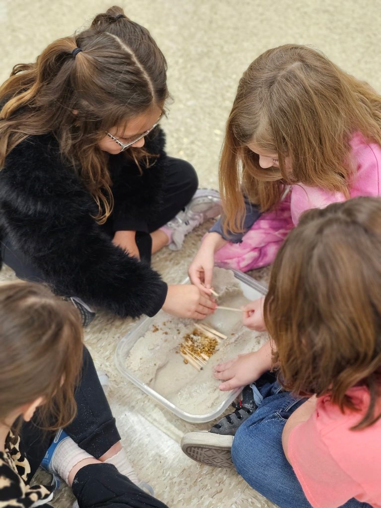Four second grade students sit on the floor around a shallow container of sand and wooden sticks, working together to build a small dam.