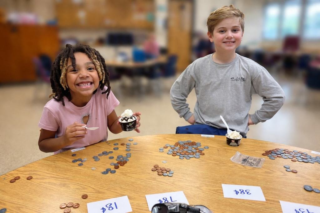 A second grade student smile while holding ice cream sundaes at a table covered with coins labeled by dollar amounts.