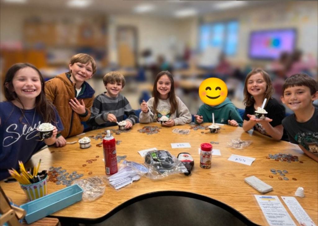 A group of smiling second grade students sit at a table holding small ice cream sundaes with coins spread out in front of them during a math activity.