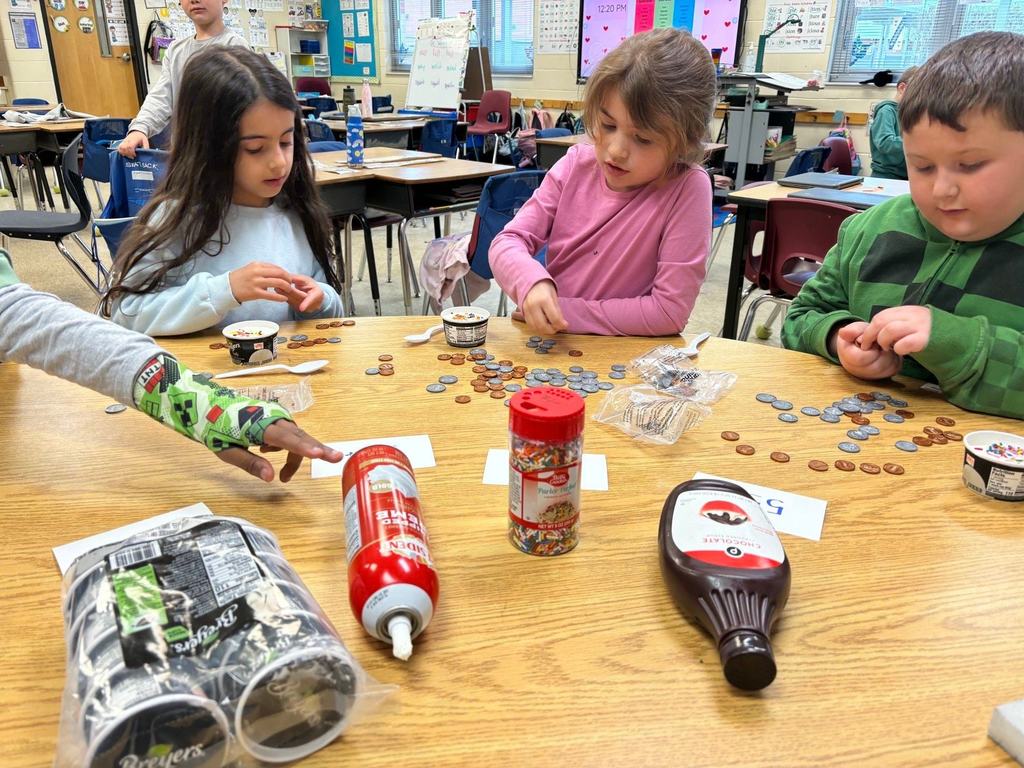 Second grade students sit around a classroom table sorting coins and counting money while ice cream sundae supplies are placed in the center.
