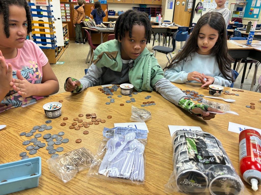 Second grade students sit at a classroom table counting coins and reviewing prices as part of a hands-on money lesson.