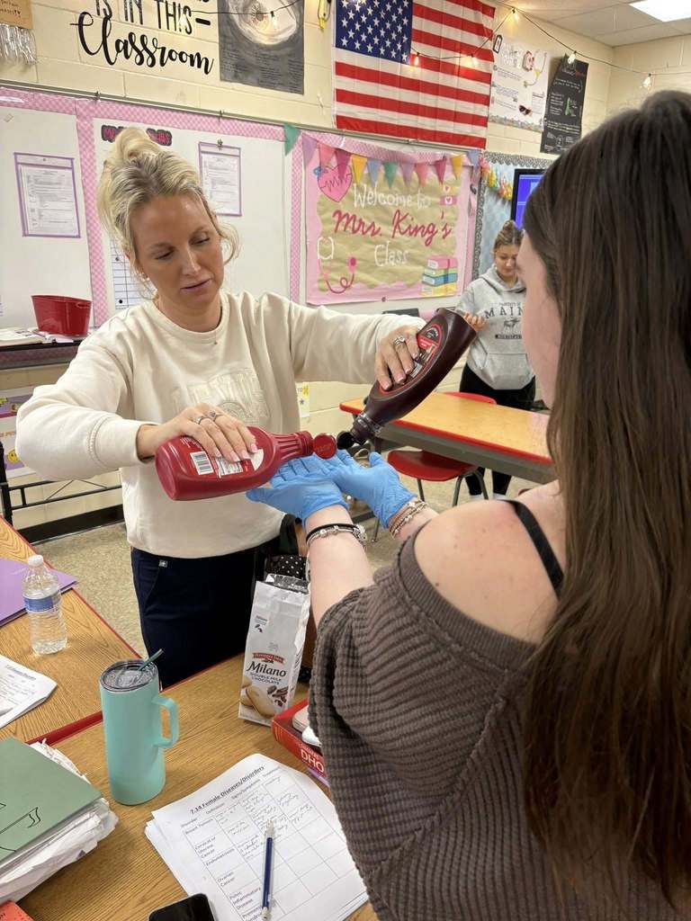 An instructor pours syrup onto a student’s gloved hands while explaining proper glove removal techniques.