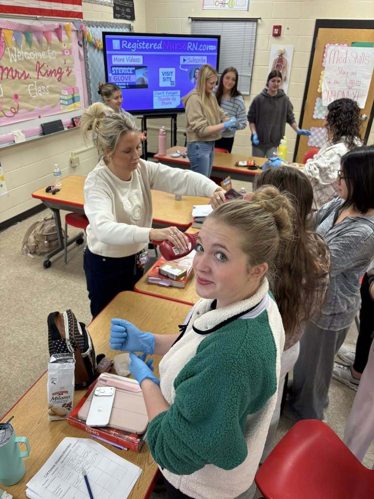 CNA students stand around classroom tables wearing blue gloves as an instructor demonstrates a hands-on infection control activity.