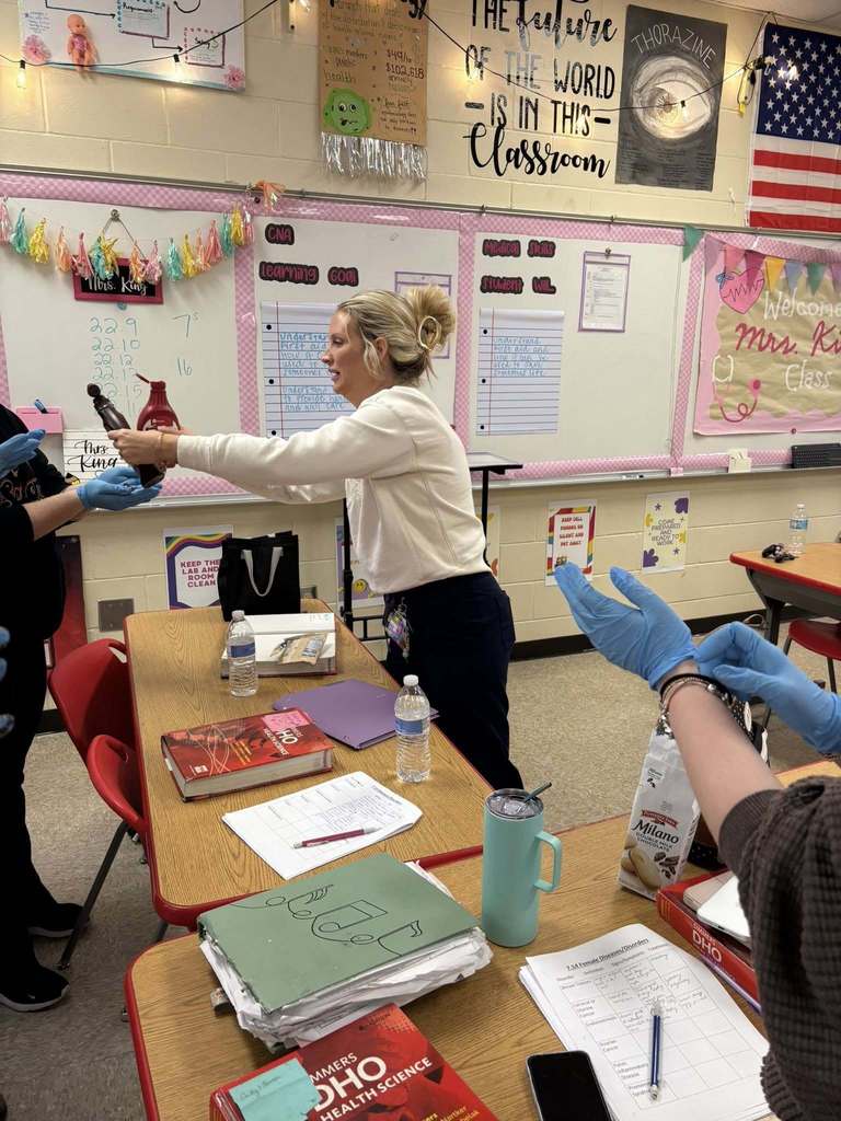 CNA students practice removing contaminated gloves while standing around a classroom table during a skills lesson.