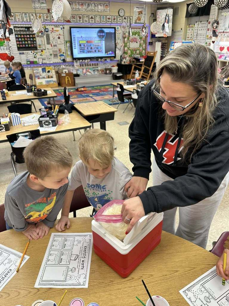 A teacher assists kindergarten students as they observe and discuss a blubber insulation demonstration in a decorated elementary classroom focused on Arctic learning.