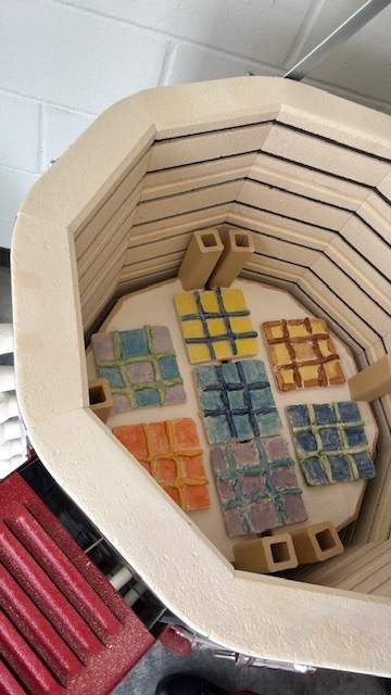 Several ceramic Tic-Tac-Toe boards arranged inside a kiln, ready for firing, showing a variety of glaze colors and grid designs.
