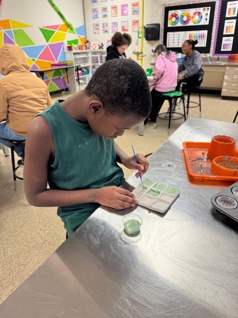 A student concentrates on painting green glaze onto a ceramic Tic-Tac-Toe board while seated at a stainless steel art table.