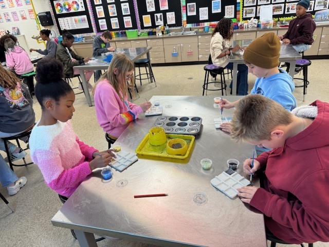 Fifth grade students seated at art tables painting clay Tic-Tac-Toe boards, focusing carefully as they apply glaze with small brushes in an art classroom.