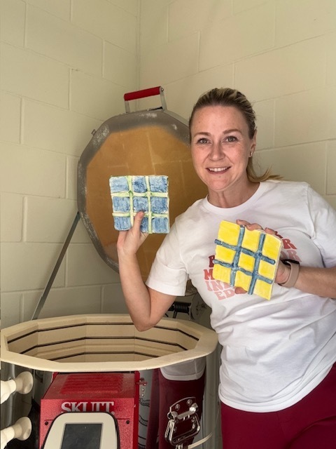 An art teacher smiles while holding two finished ceramic Tic-Tac-Toe boards next to an open kiln inside the art classroom.