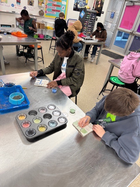 Multiple students work independently at art tables, painting and glazing ceramic Tic-Tac-Toe boards using trays of colored glaze.