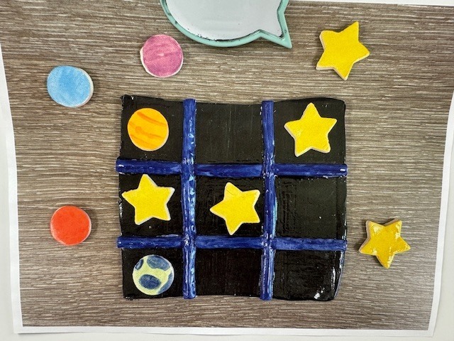 A finished ceramic Tic-Tac-Toe board with a black background and blue grid lines, surrounded by small star- and circle-shaped game pieces painted in bright colors.