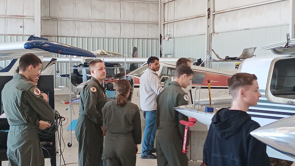 Students in aviation uniforms stand inside an aircraft hangar near small airplanes while an instructor explains components of an aircraft. Tools and maintenance equipment are visible around the planes.