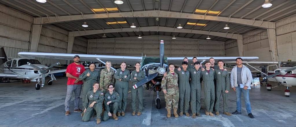 A group photo of aviation students and instructors standing in front of a small aircraft inside a hangar. Students wear matching green uniforms and pose together for the picture.