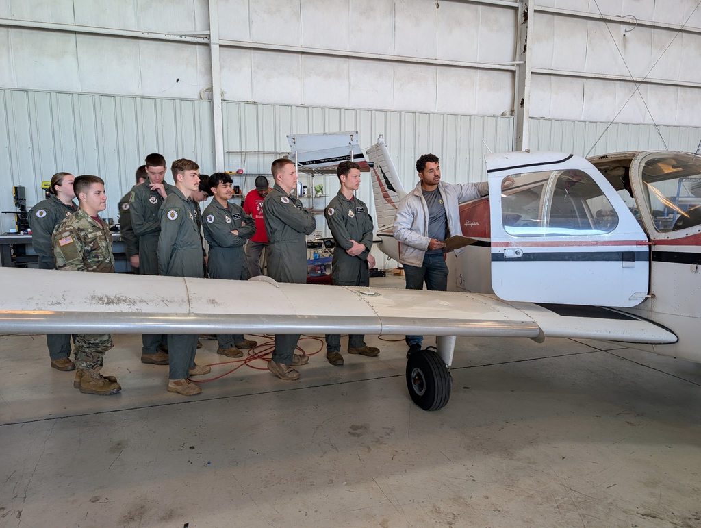 An instructor points to the cockpit area of a small airplane inside a hangar while students in aviation uniforms stand nearby listening. The aircraft wing and landing gear are visible in the foreground.