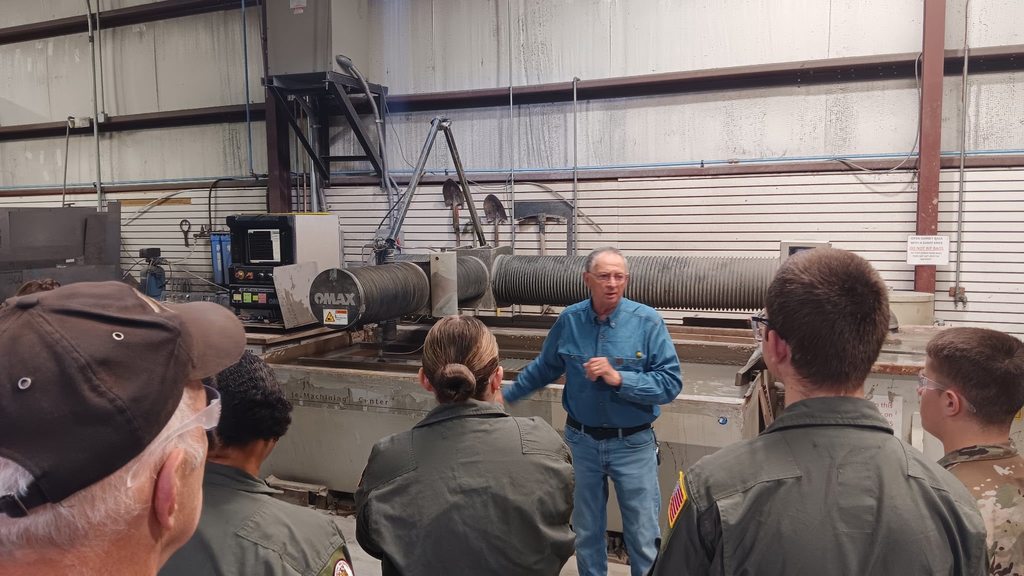 An instructor speaks to students inside an industrial machining facility. Large manufacturing equipment and a waterjet cutting machine are visible behind him as students listen attentively.