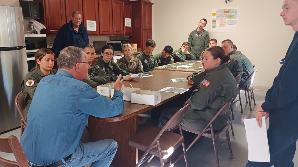 Students in green aviation-style uniforms sit around a table listening to an instructor speak in a classroom with wooden cabinets and appliances in the background. Papers are spread across the table as students focus on the discussion.