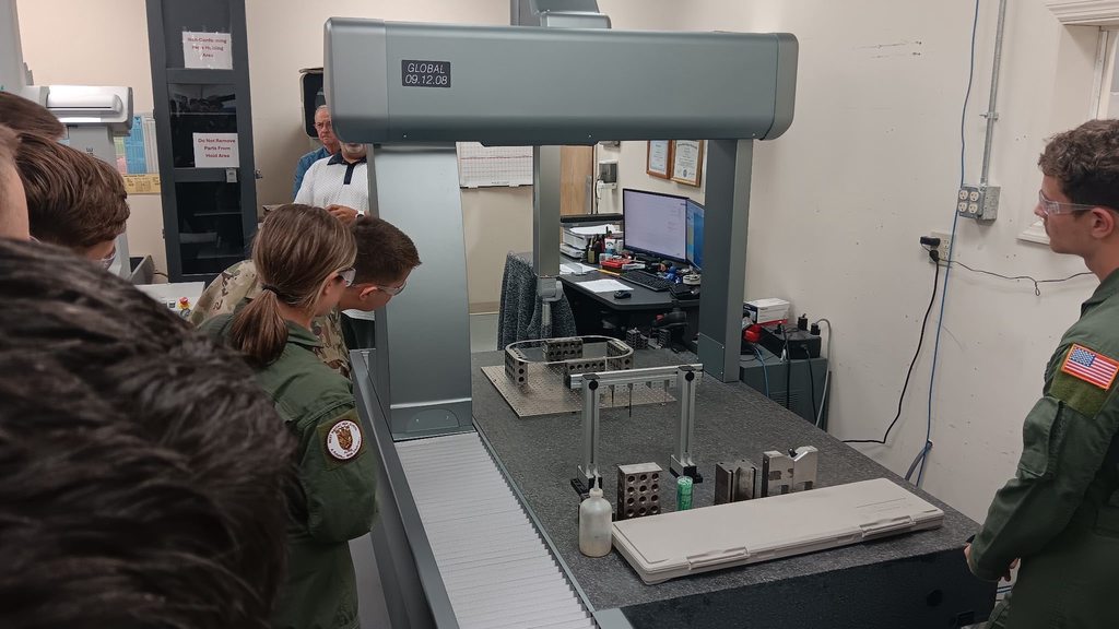 Students wearing aviation uniforms observe a large coordinate measuring machine in a lab setting while an instructor stands in the background. Metal parts and measuring tools are arranged on the inspection table.