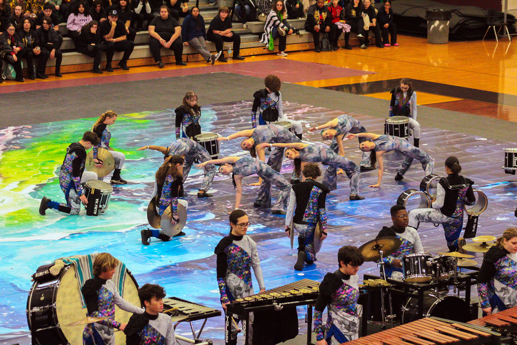 Yulee High School percussion ensemble performs a competition show on a colorful floor tarp inside a gymnasium while an audience watches from the bleachers.