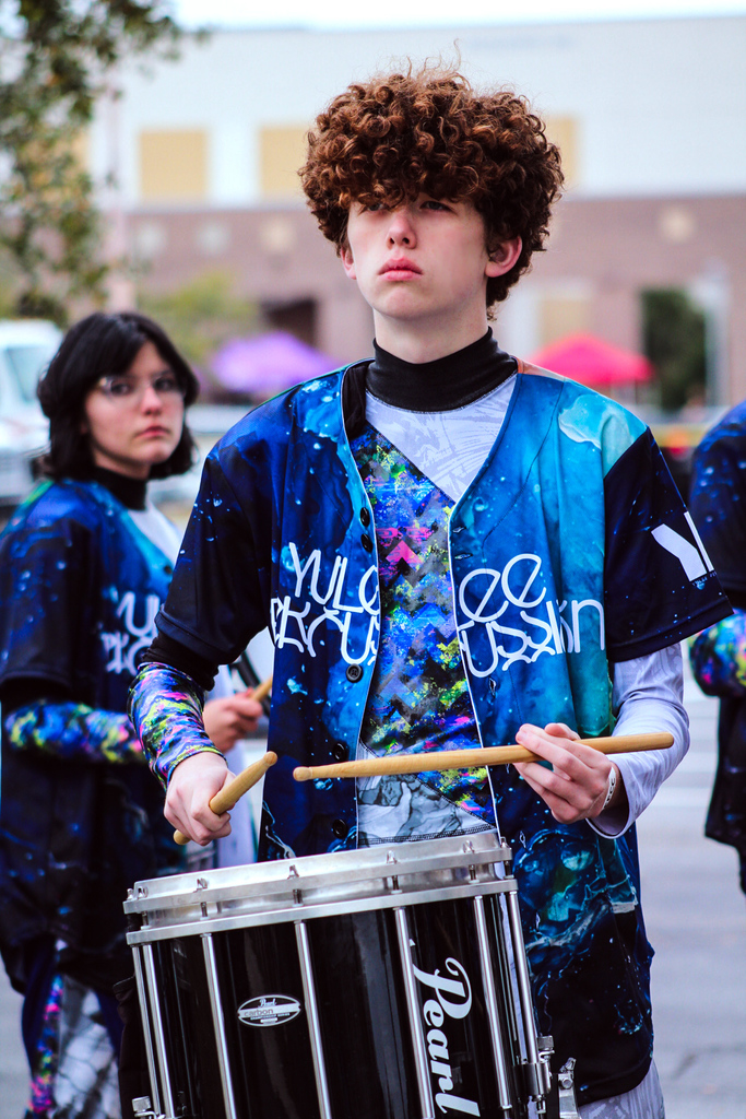 Yulee High School student in blue and teal band uniform plays a snare drum outdoors during competition
