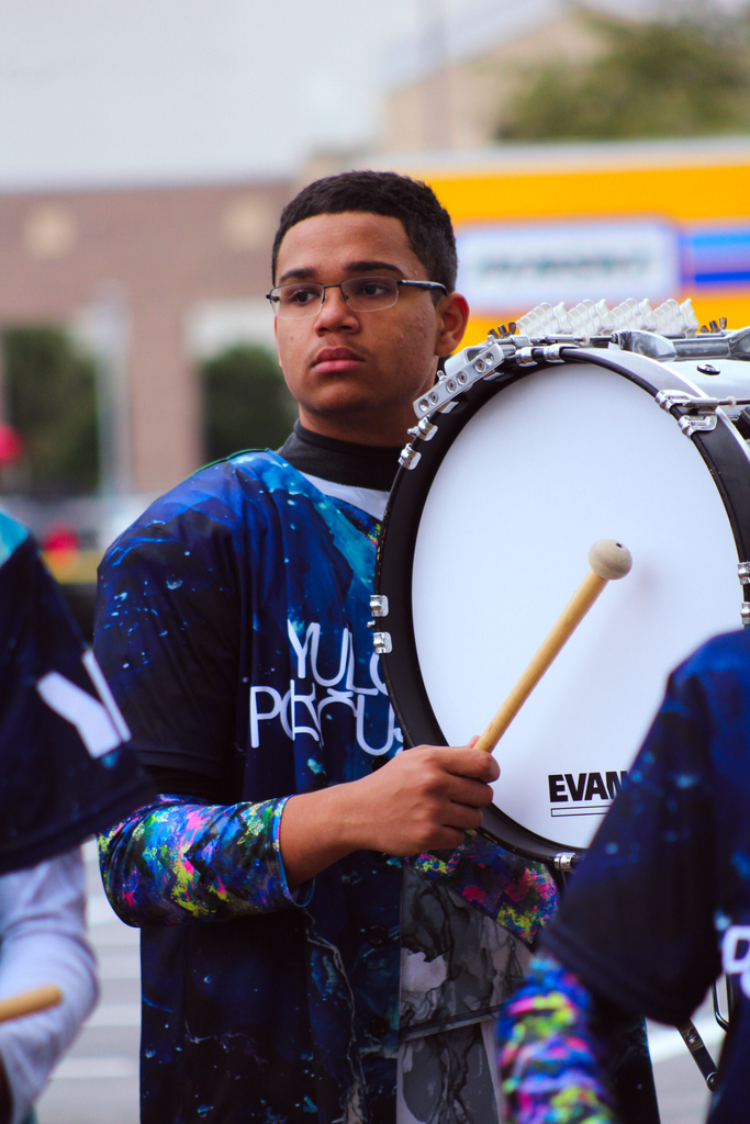 Yulee High School percussion student carries and plays a bass drum while standing in formation outdoors.