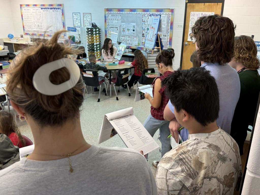 Yulee High School FFEA members stand at the back of a primary classroom observing a teacher leading a small group lesson with young students seated at a table.