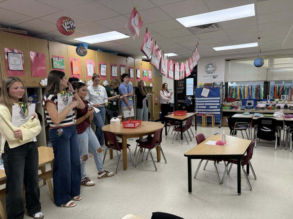 Several Yulee High School students stand along the wall of a Kindergarten classroom observing instruction while holding observation forms and notes.