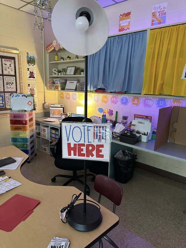 A “Vote Here” sign displayed on a stand in a colorful kindergarten classroom with alphabet borders, storage bins, and a cardboard voting booth in the background.