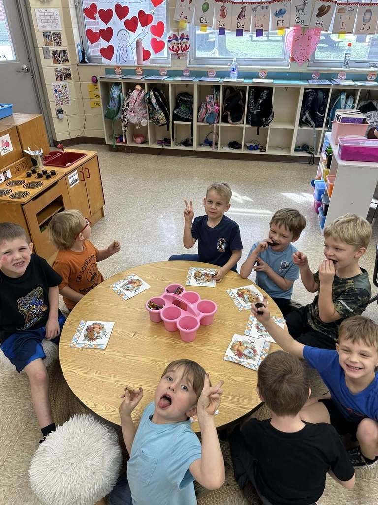 Kindergarten students sit around a classroom table eating cookies and smiling at the camera, with individual napkins and a shared container of candy in the center; cubbies and alphabet cards are visible in the background.