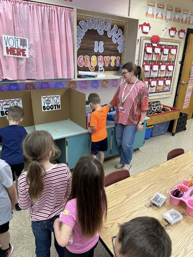 A teacher stands beside a cardboard voting booth labeled “Voting Booth” while a kindergarten student prepares to cast a ballot; a classroom display reads “Kindergarten is Groovy” and other students wait nearby.