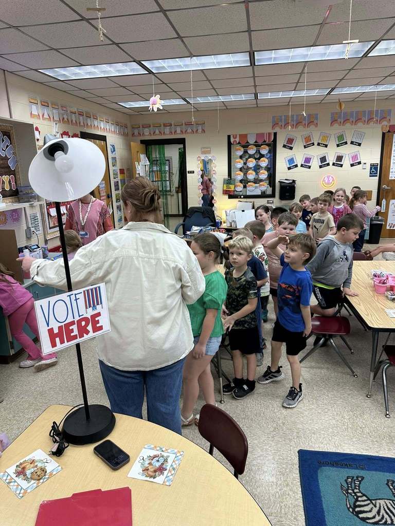 Kindergarten students form a line to vote in a classroom mock election; a “Vote Here” sign on a stand marks the voting area while a teacher assists students at the booth.