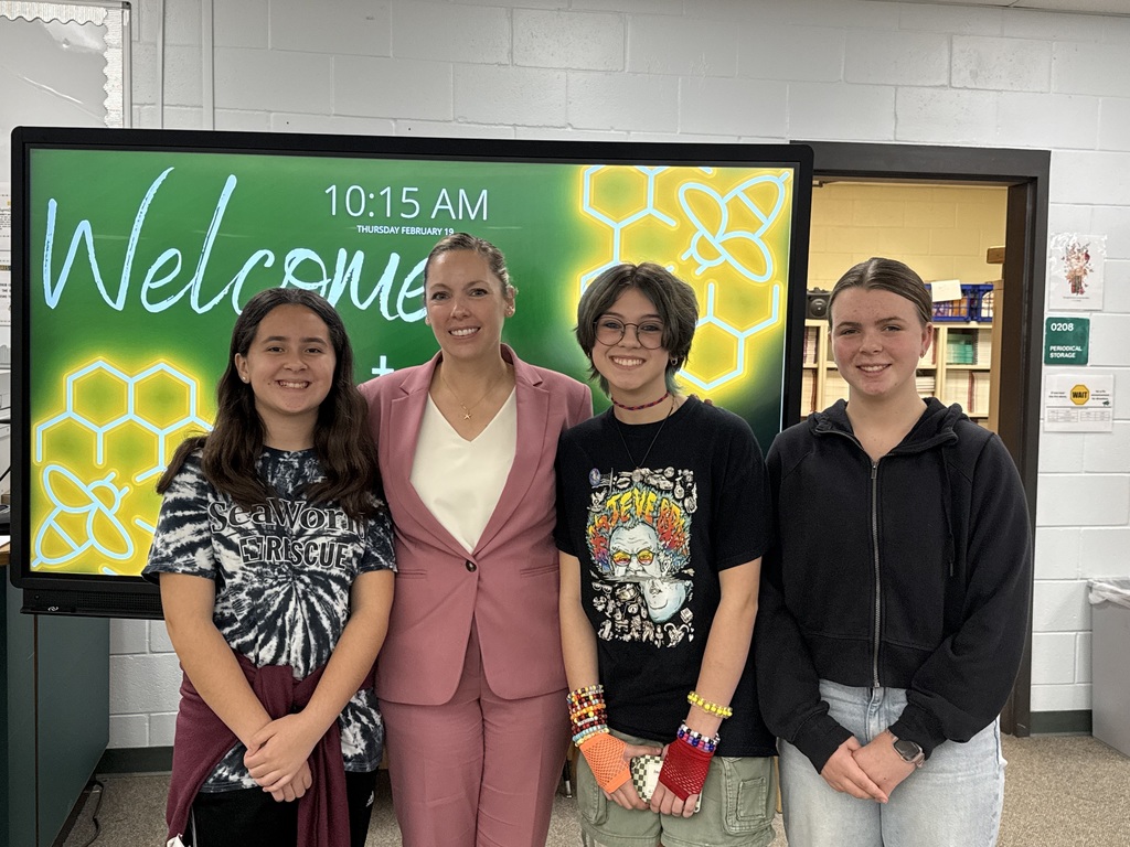Dr. Middleton, the school principal, is posing and smiling with three student winners of the 4-H speech contest. They are standing in front of a large TV with a green background and yellow bee hives on it. The screen tells the time and says Welcome. 