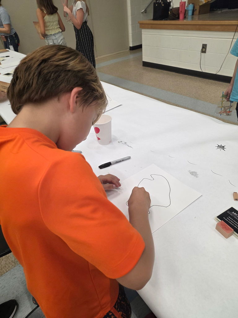 A young student in an orange shirt leaning over a large sheet of white paper, carefully drawing with a marker during a Storybook STEAM Night activity.