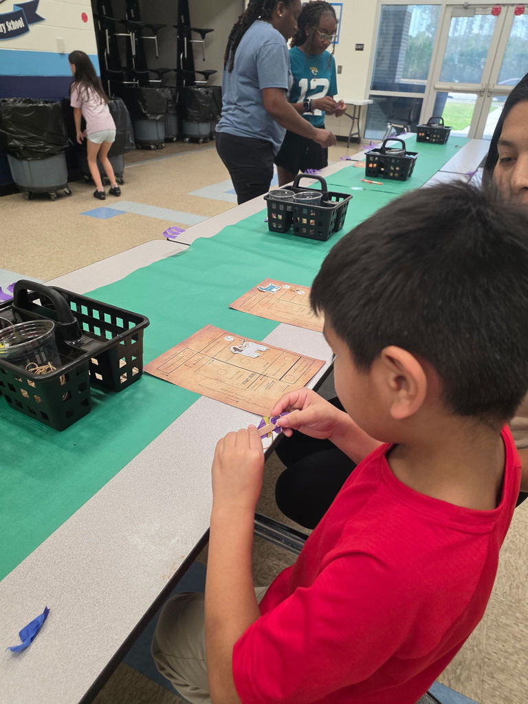 A young boy in a red shirt focused on a STEAM project, carefully using a purple marker to draw on a piece of cardboard at a long table.
